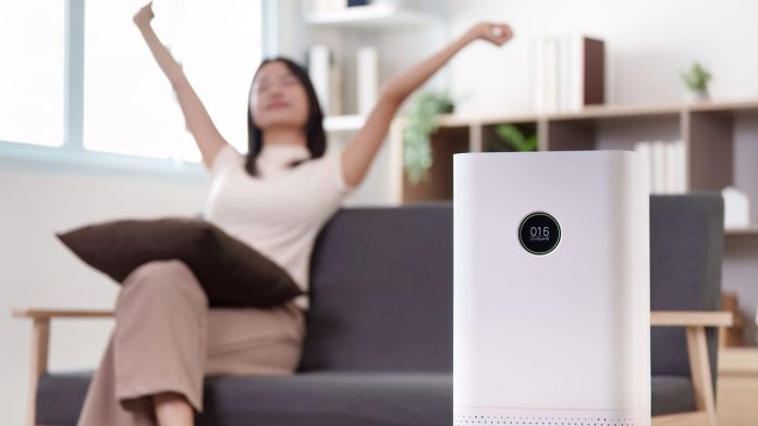 Hand-free view of a modern air purifier showing its air-quality number, with a person relaxing on the sofa behind it